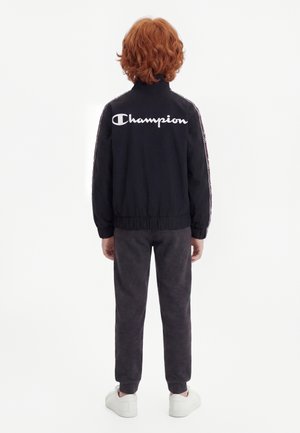 Enfant aux cheveux roux bouclés, debout de dos, portant une veste noire Champion, un pantalon sombre et des baskets blanches sur fond blanc.