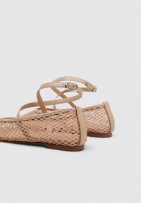 Sandales plates beiges avec un design en maille, dotées de fines brides à la cheville et d'une finition douce et texturée. La semelle est plate et marron.