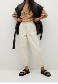 Kobieta w kwiecistej bluzce, kremowych spodniach o wysokim stanie podwiniętych przy kostkach, czarnej bezrękawnikowej kurtce skórzanej i czarnych sandałach stoi przy białej ścianie.