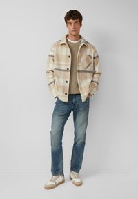 Beige kariertes Hemdjacke mit dunklen Knöpfen, getragen über einem strukturierten grauen Pullover. Blaue gerade Jeans und beige Sneaker mit hellen Akzenten.