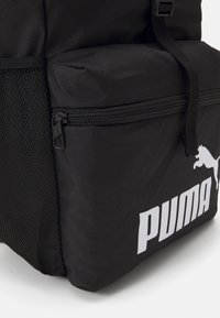 Čierny batoh s textúrovanou látkou, ktorý má zipsový priestor, sieťovanú bočnú kapsu a výrazné biele logo Puma na prednej strane.