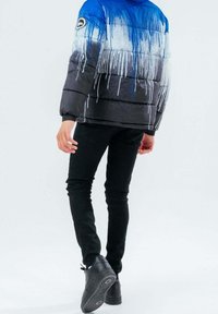 Persona con chaqueta acolchada de patrón de goteo en azul, blanco y negro, pantalones ajustados negros y zapatillas negras, caminando de espaldas sobre un fondo blanco.