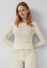 Frau mit hellbraunem Haar, die ein cremefarbenes Langarmshirt und helle Hosen trägt, steht mit den Händen hinter ihrem Rücken.