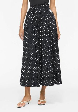 Femme portant une jupe midi noire à pois blancs, associée à des sandales blanches à talons bas et à bouts ouverts, debout sur fond blanc.