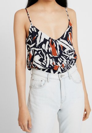 Kvinde iført ærmeløs top med abstrakt mønster i sort, hvid og rust, med tynde stropper, stoppet ned i højttaljede lyseblå jeans.