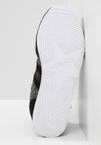 Baskets noires avec une tige en mesh texturé et des rayures. Semelle en caoutchouc blanche avec un motif d'étoiles et le logo "Versace Jeans" en relief.