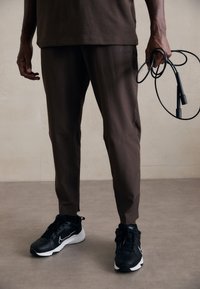 Pantalons de sport marron avec poches latérales, associés à des baskets Nike noires avec un swoosh blanc ; une corde à sauter est tenue dans la main gauche.