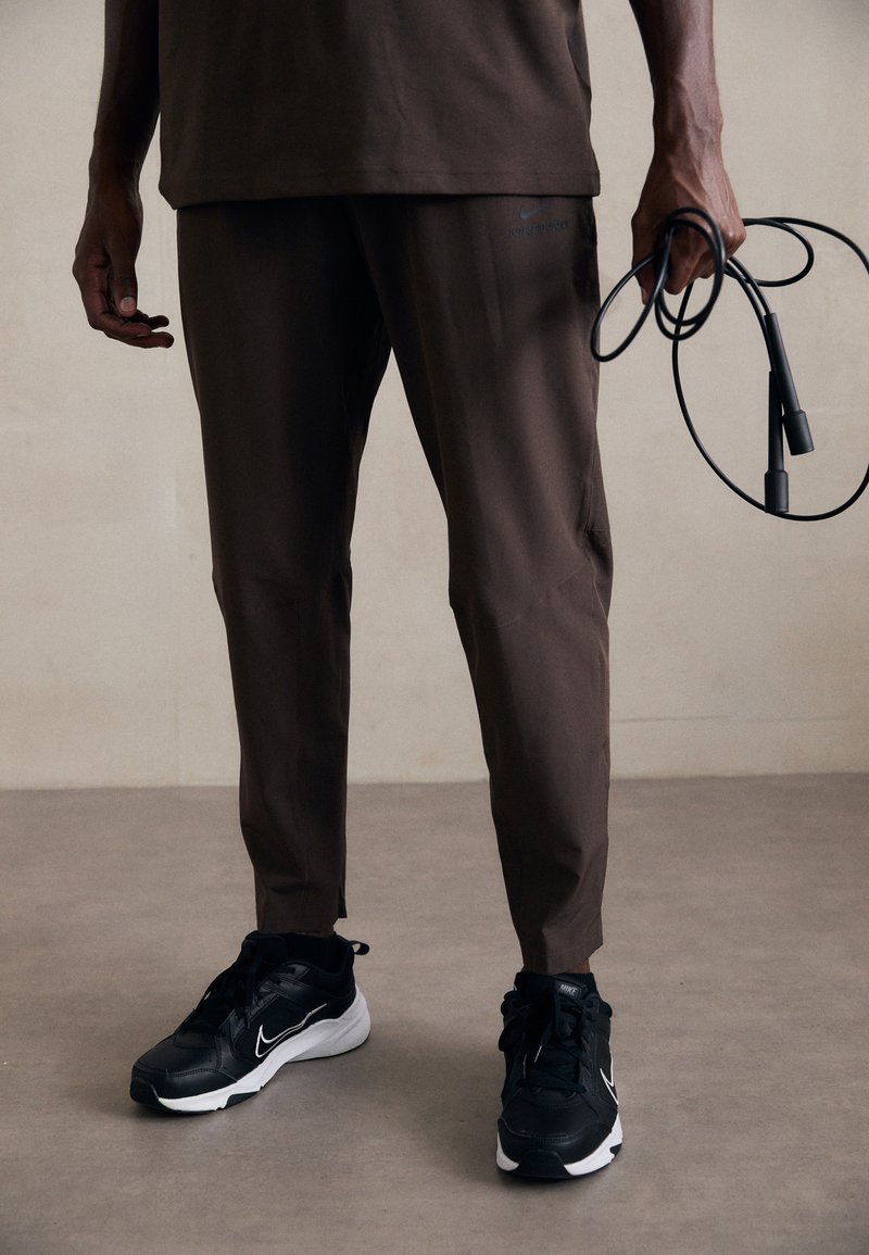 Brown athletic pants with side pockets, paired with black Nike sneakers featuring a white swoosh; a jump rope is held in the left hand.
