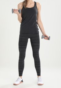 Grå ärmlös linne, randiga svarta leggings och vita sportskor med rosa detaljer. Håller ljusgrå hantlar i varje hand.