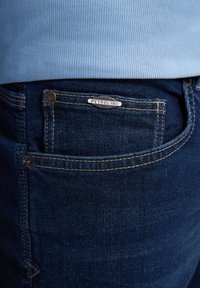 Jeans de mezclilla azul oscuro con detalle en el bolsillo trasero, que presenta una etiqueta plateada de "PETROLIND" y costuras en contraste. Textura suave.