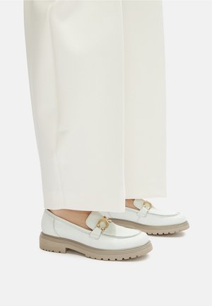 Světle zelené kožené mokasíny s kulatou špičkou, zlatými kovovými doplňky a masivní béžovou podrážkou. Kombinovány s bílými kalhotami s širokými nohavicemi.