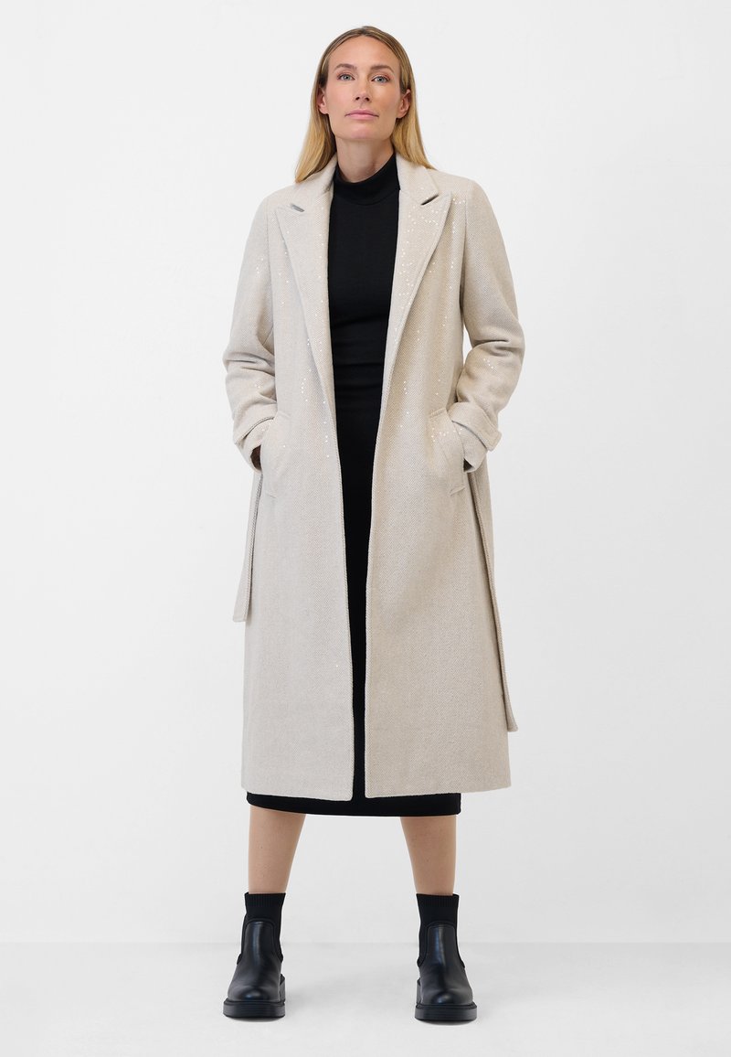 Beige long coat with a textured fabric, featuring a notched collar and side pockets, worn over a black dress and ankle boots.