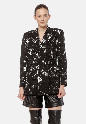 Kobieta ubrana w czarną, podwójnie zapiętą marynarkę z białym nadrukiem w plamy, czarne skórzane szorty i czarne kozaki sięgające uda, stojąca na białym tle.