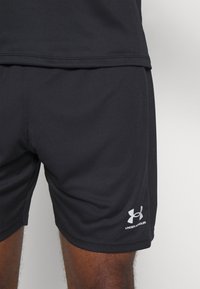 Mustat urheilushortsit, jotka ovat valmistettu kevyestä kankaasta ja joilla on sileä pinta. Alareunassa on brodeerattu Under Armour -logo.