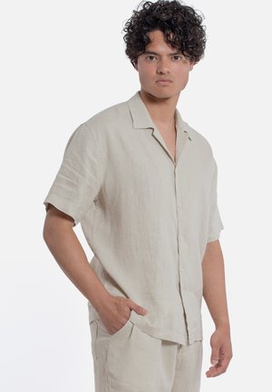 Giovane uomo con capelli ricci che indossa una camicia beige chiaro a maniche corte con bottoni e pantaloni abbinati, mano in tasca, espressione neutra.