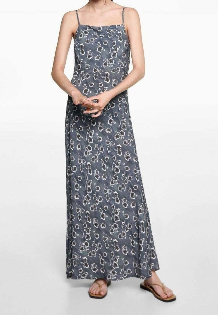 Femme portant une longue robe gris foncé à motif floral avec de fines bretelles, debout les mains jointes, et des sandales marron sur fond uni.