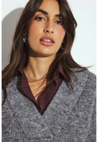 Moteris su ilgais tamsiais plaukais, dėvinti sidabrines auskarų žiedus, bordo spalvos marškinėlius su apykakle ir pilką tekstūruotą megztinį prieš vienodą baltą foną.