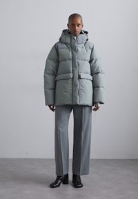 Light grey puffer jacket with a hood, featuring large pockets and a quilted texture. Paired with gray trousers and black heeled boots.