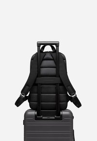 Sac à dos noir avec texture matelassée et rembourrée, attaché à une valise trolley grise avec poignée extensible. Les bretelles sont réglables.