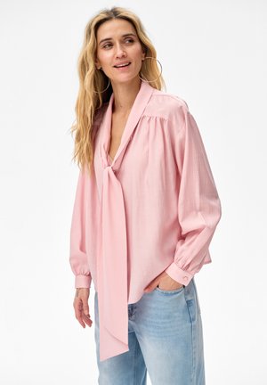 Vrouw met lang blond haar draagt grote oorringen, een loszittende roze blouse met een halsstrik en lichtblauwe jeans, staand met één hand in de zak.