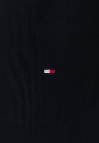 Tessuto a maglia nero con un piccolo logo ricamato in rosso, bianco e blu. La texture è liscia con un sottile motivo a coste.