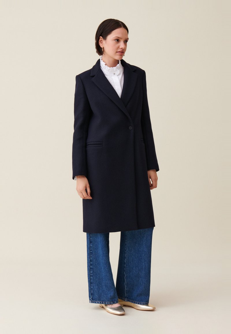 Donna che indossa un cappotto singolo blu navy scuro con un bottone, una blusa bianca con colletto arruffato, jeans a gamba larga blu e ballerine oro metalliche, in piedi contro un muro semplice.