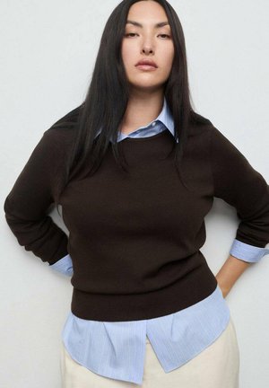 Una joven con el pelo negro largo se encuentra de pie con las manos en las caderas, vistiendo un suéter marrón oscuro sobre una camisa a rayas azul y pantalones claros.