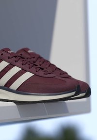 Baskets Adidas couleur bordeaux avec des bandes blanches, semelles noires et lacets, présentées sur une surface blanche avec un arrière-plan flou.