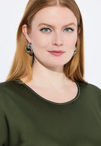 Blusa verde oliva con escote redondeado y costuras claras. La persona que la lleva puesta tiene grandes pendientes circulares de plata y el cabello liso.