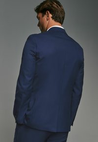  marineblaues Sakko aus strukturiertem Stoff. Verfügt über einen Zwei-Knopf-Verschluss und eine Slim-Fit-Form. Modell von hinten gezeigt, rechter Ärmel sichtbar.