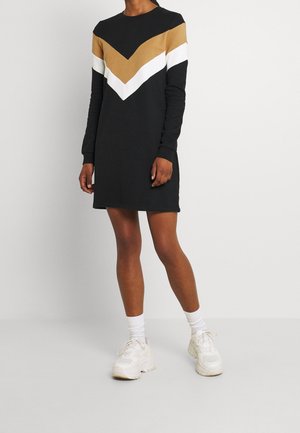 Woman wearing a black long-sleeve dress with tan and white chevron stripes, paired with white socks and chunky white sneakers.