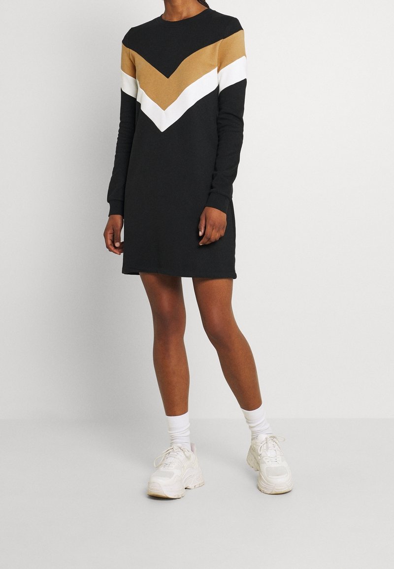 Femme portant une robe noire à manches longues avec des rayures chevrons beiges et blanches, assortie à des chaussettes blanches et des baskets blanches épaisses.
