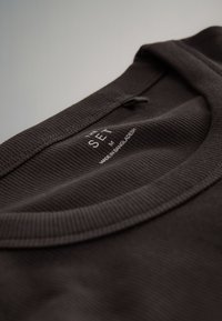 Dunkelbraunes, geripptes Baumwollshirt mit Rundhalsausschnitt. Etikett trägt die Aufschrift "THE SET M" und "MADE IN BANGLADESH." Glatte Textur.