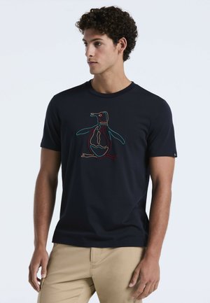 Jonge man met krullend haar, gekleed in een marineblauw T-shirt met een kleurrijke omrande pinguïnafbeelding en beige broek, kijkt opzij.