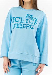 Svetlo modra majica s kapuco v sproščenem kroju, ki ima na sprednji strani velik grafični print besede "ICEBERG" v kontrastni modri barvi.