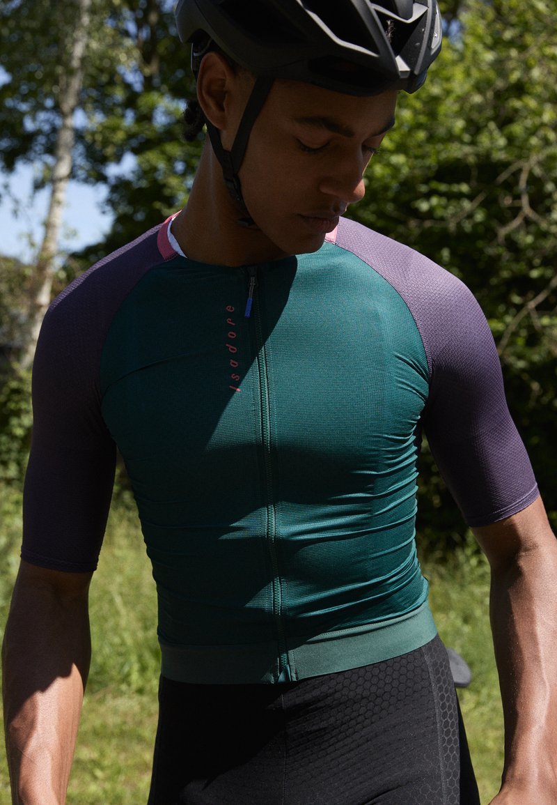 Kortärmad cykeltröja i mörkgrön med lila accenter, med en dragkedja framtill och texturerad tyg. Integrerad med figurnära svarta cykelshorts.