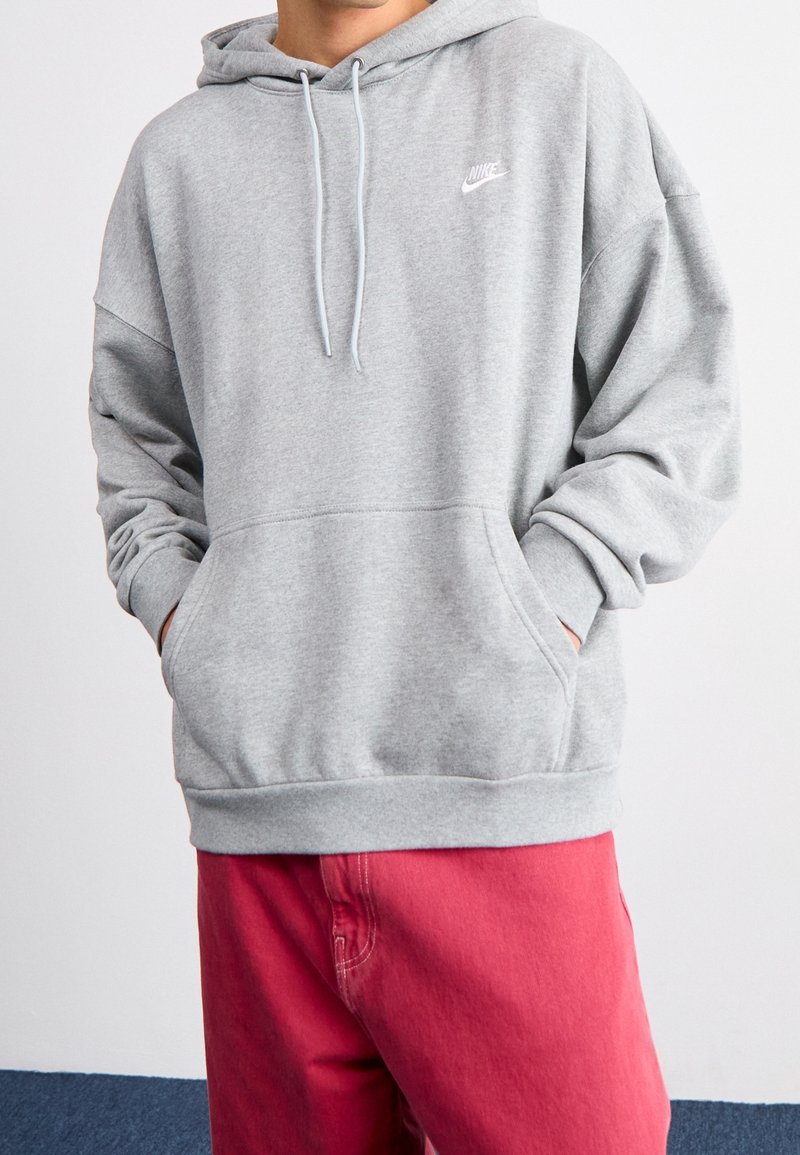 Personne portant un sweat à capuche Nike gris clair avec les mains dans la poche avant et un pantalon rouge vif, debout devant un fond blanc uni.