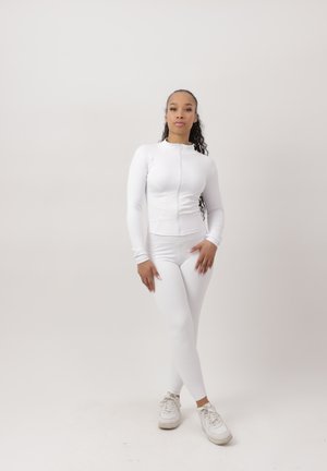 Donna con lunghi capelli ricci che indossa una giacca bianca con cerniera, leggings bianchi e scarpe da ginnastica bianche, in piedi contro uno sfondo chiaro e semplice.