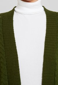 Cardigan en maille côtelée verte à ouverture frontale, associé à un col roulé blanc. Tissu texturé avec une apparence douce et confortable.