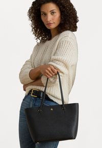 Saco tote preto com uma superfície texturizada, alças duplas e detalhes em dourado. Detalhe do logótipo notável na frente, forma retangular.