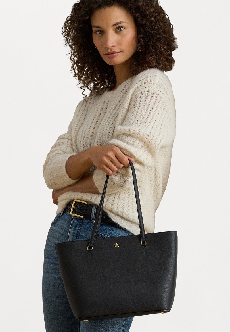 Saco tote preto com uma superfície texturizada, alças duplas e detalhes em dourado. Detalhe do logótipo notável na frente, forma retangular.