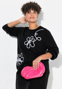 Schwarzer Pullover mit weißen abstrakten Designs, kombiniert mit einer rosa Clutch. Das Model hat lockiges Haar und trägt Ohrstecker. Strukturierter Stoff.