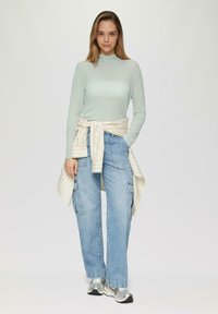 Lichtgroene langemouwen top met een opstaande hals, gecombineerd met losse blauwe cargo jeans en een roomkleurig gestreept shirt dat om de taille is geknoopt. Zilveren sneakers.