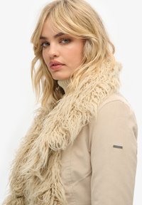 Beige textuursjaal met een harige uitstraling, gedragen over een lichtgekleurde jas. De sjaal heeft zachte, pluizige strengen voor zowel warmte als stijl.