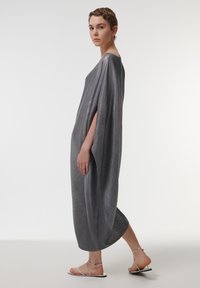 Kaftán largo y gris con un acabado texturizado, con mangas amplias y un corte holgado, combinado con sandalias de tiras.