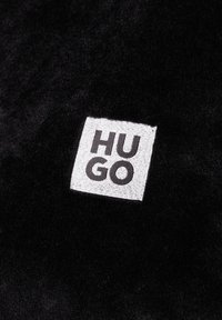 Czarna tkanina o miękkiej teksturze ma kwadratową białą łatę z napisem "HUGO" w grubych czarnych literach, wskazującą na markę.