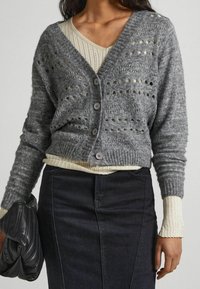 Grå stickad cardigan med V-ringning, med öppet mönster, fem knappar, ribbade ärmslut och en mjuk textur över en ljus topp och mörk kjol.