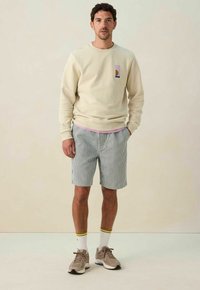 Sweatshirt beige avec un design graphique sur la poche, associé à un short rayé bleu et blanc. Baskets grises et chaussettes blanches avec des accents jaunes.