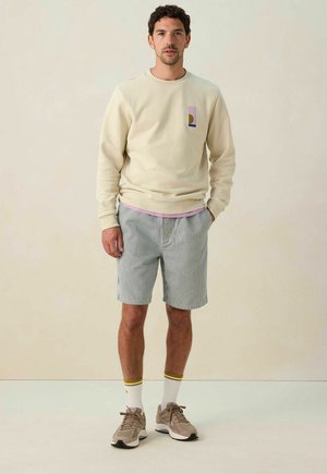 Sweatshirt beige avec un design graphique sur la poche, associé à un short rayé bleu et blanc. Baskets grises et chaussettes blanches avec des accents jaunes.