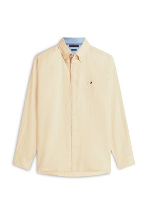 Beige pitkähihaiset napilliset paita, jossa on napitettu kaulus ja pieni logo vasemmassa rinnassa valkoisella taustalla.